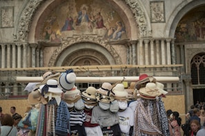 A variety of stylish hats are displayed on a stand in front of an intricately decorated architectural structure. The building features ornate carvings and religious frescoes above a large archway. Several people are gathered nearby, looking at the items or walking past.