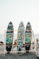 Three colorful surfboards with vibrant graffiti-style artwork are displayed on a wooden rack. Each board has a unique design featuring abstract and cartoonish images against the backdrop of a sandy beach with people in the distance and the ocean visible under a clear sky.