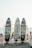 Three colorful surfboards with vibrant graffiti-style artwork are displayed on a wooden rack. Each board has a unique design featuring abstract and cartoonish images against the backdrop of a sandy beach with people in the distance and the ocean visible under a clear sky.