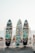 Three colorful surfboards with vibrant graffiti-style artwork are displayed on a wooden rack. Each board has a unique design featuring abstract and cartoonish images against the backdrop of a sandy beach with people in the distance and the ocean visible under a clear sky.