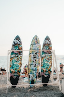 Three colorful surfboards with vibrant graffiti-style artwork are displayed on a wooden rack. Each board has a unique design featuring abstract and cartoonish images against the backdrop of a sandy beach with people in the distance and the ocean visible under a clear sky.