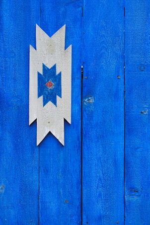 A bright blue wooden wall featuring a geometric wooden decoration in the center. The decoration is an arrow-like shape with a blue star design in the middle and a small diamond in red at the center.