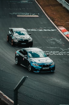 Two sports cars are racing on a paved track with graffiti and markings. The cars appear to be in motion on a winding part of the track, and there's a fence in the foreground. The scene is set in an outdoor location with a partly grassy area visible along the edges of the track.