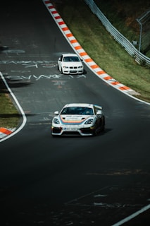 Two sports cars are racing on a track flanked by grassy banks and safety barriers. The road surface has various markings and graffiti. One car is a Porsche with a racing livery in the foreground, while a white BMW follows behind.
