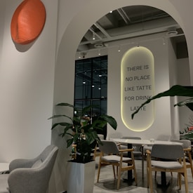 A modern indoor cafe space with sleek furnishings, featuring a large white arch leading to a seating area. Grey chairs and a light wooden table are surrounded by potted green plants. A wall displays the statement 'THERE IS NO PLACE LIKE TATTE FOR DRINK LATTE' in black lettering, highlighted with soft backlighting. An orange textured wall feature adds a pop of color.