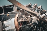 Close-up view of a vintage airplane engine featuring an exposed radial engine with intricate metallic components, bolts, and a large propeller. The metallic surfaces display a range of textures including polished and aged metal, giving a sense of craftsmanship and engineering complexity. Sunlight creates a warm cast on the engine, highlighting the details.