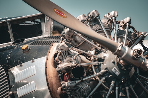 Close-up view of a vintage airplane engine featuring an exposed radial engine with intricate metallic components, bolts, and a large propeller. The metallic surfaces display a range of textures including polished and aged metal, giving a sense of craftsmanship and engineering complexity. Sunlight creates a warm cast on the engine, highlighting the details.