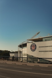 A modern stadium building with a large emblem of 'Toulouse Football Club' is seen against a clear blue sky. The structure has a sleek design with metal beams and glass windows, located next to a road with protective railings. Trees and greenery are visible in the background, adding a natural element to the urban scene.