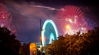 A vibrant display of fireworks illuminates the night sky over a glowing Ferris wheel. The scene is set in an urban environment with trees and a lit streetlamp in the foreground. Red and pink fireworks explode against a dark sky, while the Ferris wheel casts a turquoise glow. A promotional poster on a nearby building adds a splash of color.