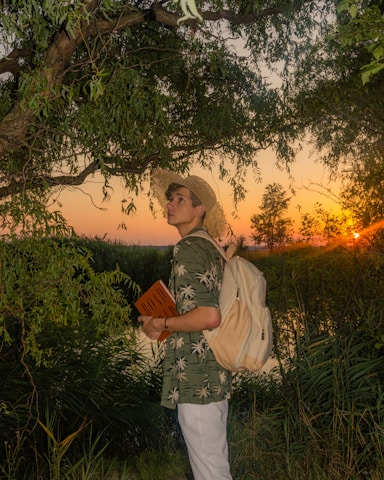 A person wearing a straw hat and a green shirt with palm tree patterns stands near a tree, holding a book titled 'ROAD'. The person has a backpack and gazes into the distance. The background features a vibrant sunset with the sun partially visible, surrounded by lush greenery.