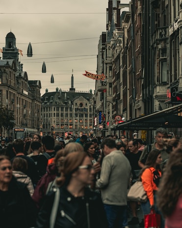 A bustling city street filled with people, with tall historic buildings lining the sides. The street has a sign for a casino and other shops. Overhead, there are suspended lights, and in the distance, more architectural features can be seen.