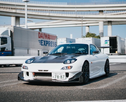 A sleek white sports car with a black hood and aerodynamic design is parked on an industrial or urban road. Surrounding the car are large trucks with visible logos, set against a backdrop of highways and blue sky.