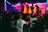 A lively outdoor concert with a band performing on stage under vibrant lighting. People are gathered in a crowd in front of the stage, some dancing in the foreground. Bright red and blue lights illuminate the scene, creating a dynamic and energetic atmosphere.