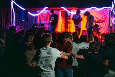 A lively outdoor concert with a band performing on stage under vibrant lighting. People are gathered in a crowd in front of the stage, some dancing in the foreground. Bright red and blue lights illuminate the scene, creating a dynamic and energetic atmosphere.