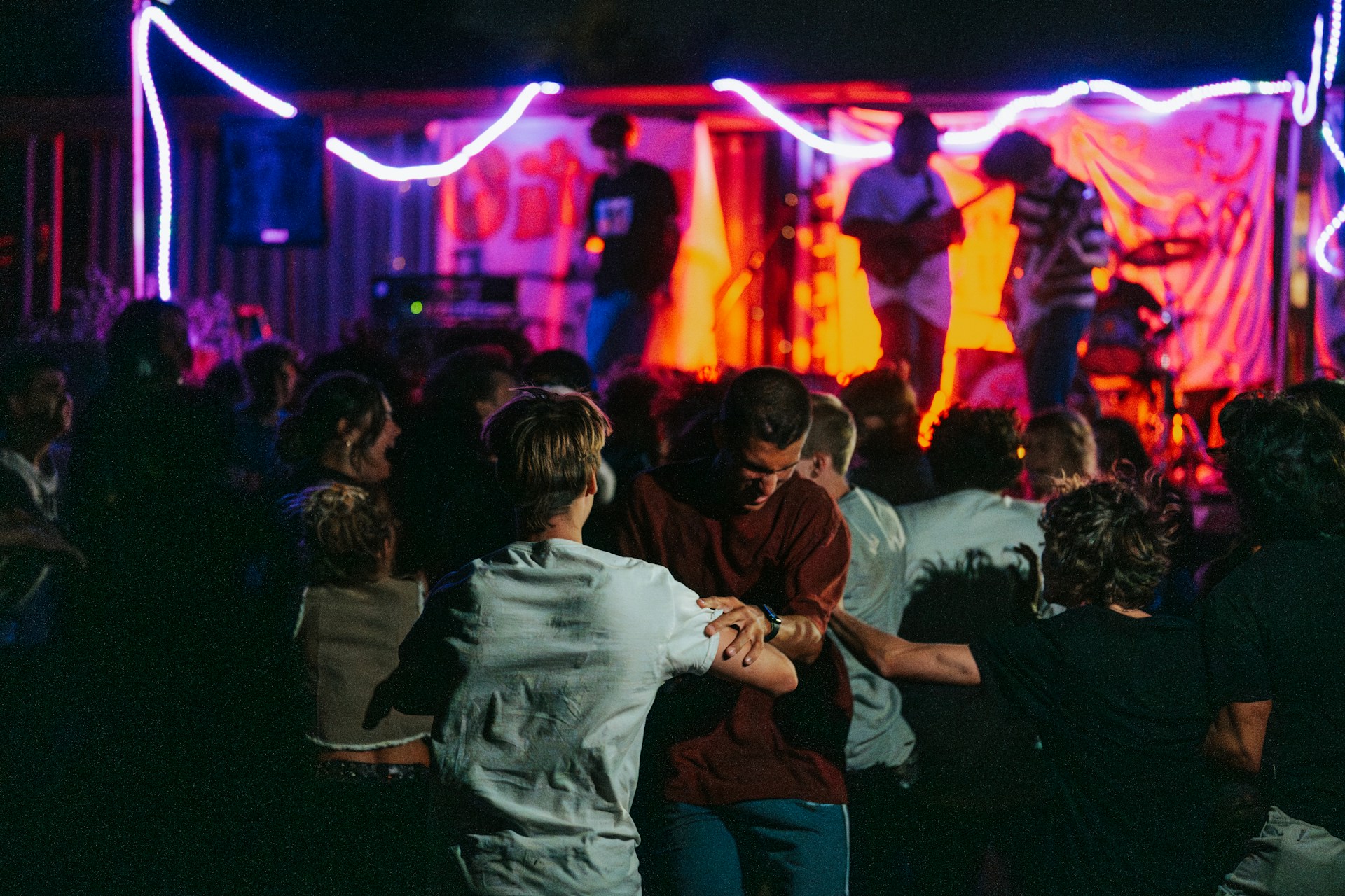 A lively outdoor concert with a band performing on stage under vibrant lighting. People are gathered in a crowd in front of the stage, some dancing in the foreground. Bright red and blue lights illuminate the scene, creating a dynamic and energetic atmosphere.