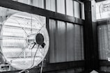 A mounted industrial fan with metal blades and a protective cage is attached to a wall inside a structure with wooden supports and corrugated metal panels. The image appears monochromatic, predominantly showcasing shades of grey and black. There is also part of a metal frame or cage visible to the right side of the image.