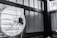 A mounted industrial fan with metal blades and a protective cage is attached to a wall inside a structure with wooden supports and corrugated metal panels. The image appears monochromatic, predominantly showcasing shades of grey and black. There is also part of a metal frame or cage visible to the right side of the image.