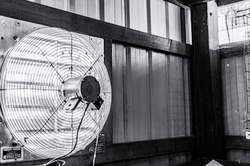 A mounted industrial fan with metal blades and a protective cage is attached to a wall inside a structure with wooden supports and corrugated metal panels. The image appears monochromatic, predominantly showcasing shades of grey and black. There is also part of a metal frame or cage visible to the right side of the image.