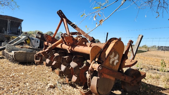 An old, rusted agricultural plow is situated on dry, grassy ground near a wire fence. The plow has a series of circular blades and appears to be unused for some time. In the background, there is a partially visible structure with a corrugated metal roof, surrounded by sparse vegetation.