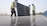 Workers wearing safety gear are installing solar panels on a rooftop. They are dressed in safety vests and helmets, and are handling a large solar panel as part of a series of panels. The background includes a clear sky and some infrastructure.