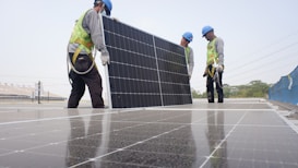 Workers wearing safety gear are installing solar panels on a rooftop. They are dressed in safety vests and helmets, and are handling a large solar panel as part of a series of panels. The background includes a clear sky and some infrastructure.