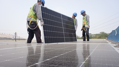 Workers wearing safety gear are installing solar panels on a rooftop. They are dressed in safety vests and helmets, and are handling a large solar panel as part of a series of panels. The background includes a clear sky and some infrastructure.