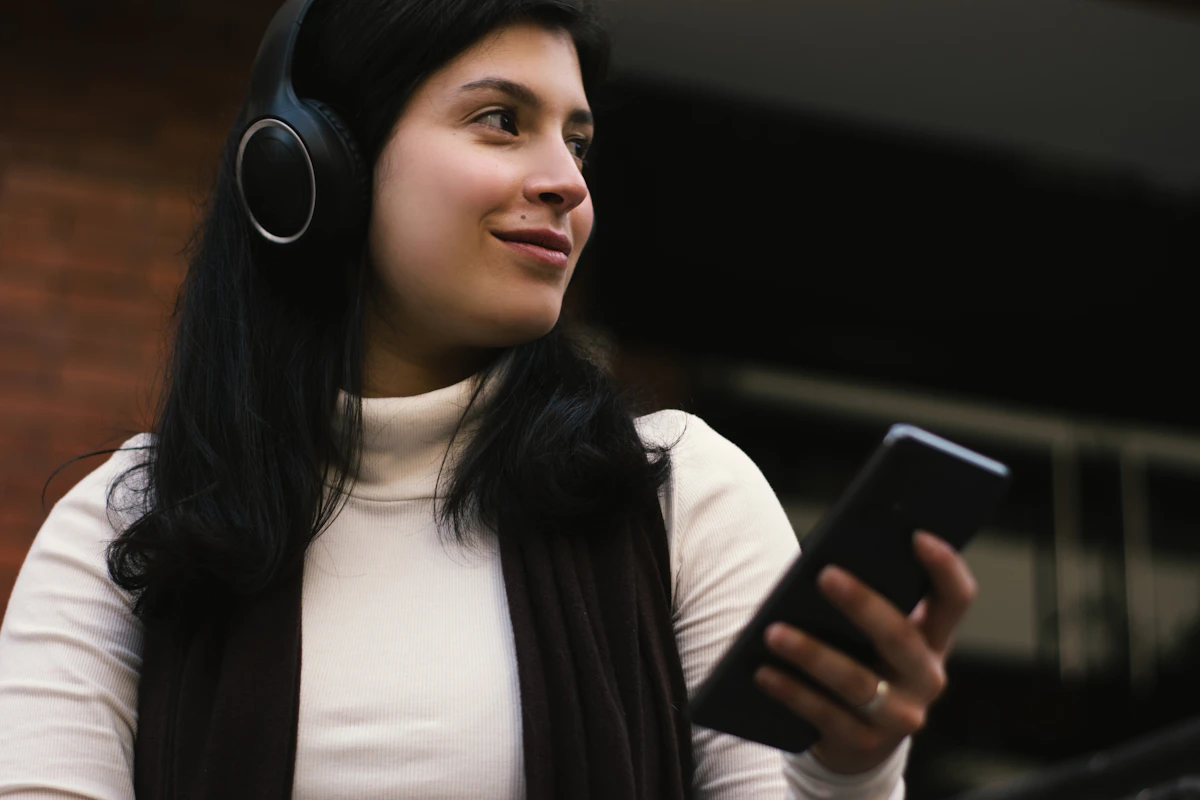 ヘッドホンをつけた黒髪の女性が、白いタートルネックの服で、手前に向けた手にスマートフォンを持ちながら、画面ではなく右の遠くのほうを見て微笑んでいる