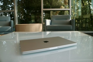 A sleek laptop is placed on a glass-top table in a modern lounge area. The background features large windows letting in natural light, with soft-cushioned armchairs and greenery visible outside.