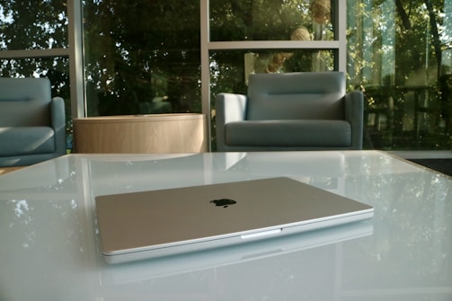 A sleek laptop is placed on a glass-top table in a modern lounge area. The background features large windows letting in natural light, with soft-cushioned armchairs and greenery visible outside.
