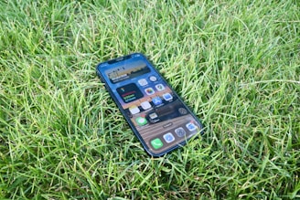 A smartphone lies face up on a lush, green grass surface. The phone's screen displays various app icons and a digital widget.