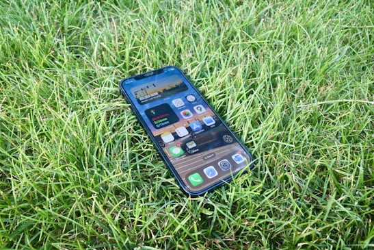 A smartphone lies face up on a lush, green grass surface. The phone's screen displays various app icons and a digital widget.