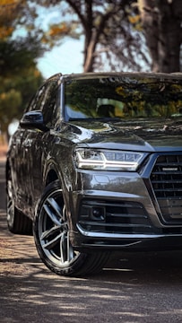 A sleek, dark-colored SUV is parked on a paved road surrounded by trees. The vehicle has a glossy finish that reflects the natural surroundings, and the intricate design of the alloy wheels is prominently visible. Sunlight dapples the scene, creating a dynamic interplay of light and shadow on the car's surface.