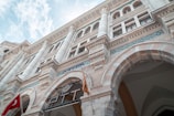 A detailed view of a grand, historical building with ornate columns, arched entryways, and intricate decorations. There are multiple stories with tall windows, and inscriptions in blue tile along the facade. A Turkish flag is prominently displayed near the entrance.