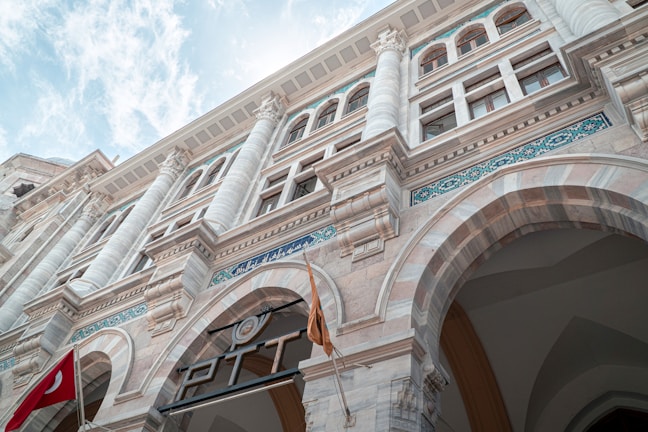 A detailed view of a grand, historical building with ornate columns, arched entryways, and intricate decorations. There are multiple stories with tall windows, and inscriptions in blue tile along the facade. A Turkish flag is prominently displayed near the entrance.