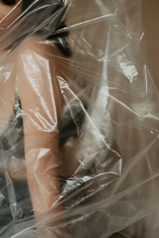 A partial view of a person's arm and shoulder wrapped in a translucent plastic sheet. The image creates a sense of texture and depth with light reflections on the plastic surface.