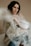 A person with dark hair and gentle expression sits, enveloped in a translucent plastic sheet. The muted background and soft lighting create a serene and contemplative atmosphere. The person's makeup is subtle, enhancing their calm demeanor.