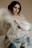 A person with dark hair and gentle expression sits, enveloped in a translucent plastic sheet. The muted background and soft lighting create a serene and contemplative atmosphere. The person's makeup is subtle, enhancing their calm demeanor.