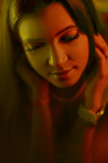 A person with long dark hair appears in a warmly lit, softly focused setting. Their eyes are closed, and they exhibit a serene expression with a slight smile. Hands gently touch the sides of their head, with a watch visible on one wrist.