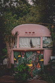 A vintage-style pink trailer with white curtains and decorative artwork at the front, surrounded by lush greenery. The artwork on the lower section features playful, colorful illustrations including leaves, flowers, and sailboats, with the words 'runaway cafe' prominently displayed.