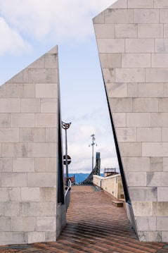 A pathway with a brick pattern stretches between two large, slightly inclining concrete structures that create a V-shaped opening. The structures have a modern, minimalist design with a light gray color. Beyond the pathway, there's a modern sculpture and a railing overlooking a scenic view of mountains under a bright sky with scattered clouds.