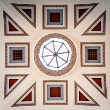 A geometrically designed ceiling featuring a central octagonal skylight surrounded by a series of square and triangular decorative patterns. The colors include soft beige, red squares in the center of the patterns, and blue-gray framing details. The design is symmetrical, giving a sense of balance and complexity.
