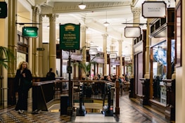 A bustling indoor shopping arcade with elegant architectural features such as tall columns, detailed moldings, and ornamental lighting. Numerous shop signs are visible, including ones for Wallace Cotton and Smith the Grocer. People are seen walking and sitting in the dining area under the warm glow of ambient lighting. The décor includes polished wood and green plants.