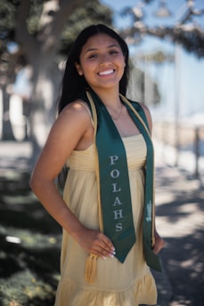 A young woman is smiling while wearing a green graduation stole with 'POLAHS' written on it. She is standing outdoors with trees and a blurred background, wearing a light yellow dress.