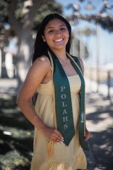 A young woman is smiling while wearing a green graduation stole with 'POLAHS' written on it. She is standing outdoors with trees and a blurred background, wearing a light yellow dress.