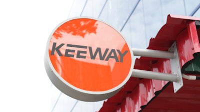 A circular sign with a bright orange background features bold black letters spelling out 'Keeway'. The sign is attached to a structure with multiple metallic rods and a red overhang, set against a reflective glass building facade.