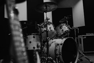 A black and white photograph of a drum set positioned in a room, with a blurred guitar in the foreground and an amplifier in the background. The lighting creates a moody and atmospheric environment.