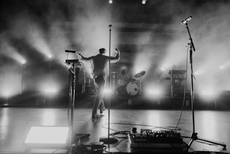 A musician in a dynamic stance is performing on a stage enveloped in dramatic lighting and mist. The scene features illuminated drums and multiple microphones on stands, enhancing the concert atmosphere.