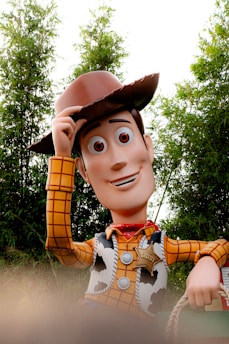 A cartoonish cowboy character stands outdoors, tipping his brown hat with one hand. He wears a yellow plaid shirt, a cowhide vest, and a red bandana. The character has large, expressive eyes and a smile. Behind him, green foliage provides a natural backdrop.