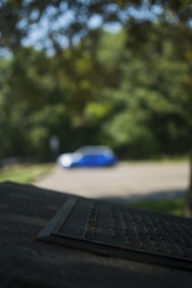 A blurred image displaying a metallic plaque in the foreground with engraved text, partially in focus. In the background, there is a blue and white object, possibly a car, situated in a parking area surrounded by lush greenery and trees.