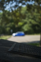 A blurred image displaying a metallic plaque in the foreground with engraved text, partially in focus. In the background, there is a blue and white object, possibly a car, situated in a parking area surrounded by lush greenery and trees.